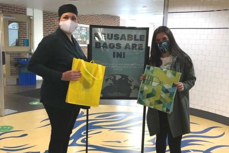 Two people hold reusable bags in front of a sign
