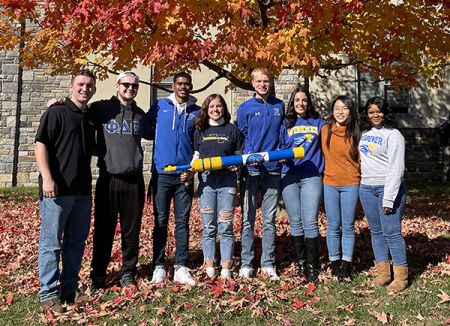Eight students pose for a photo with the center student holding a T-shirt launcher.