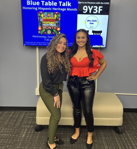 Students Maritza Garcia and Ariana Hurtado-Day stand in front of a screen advertising a Blue Table Talk