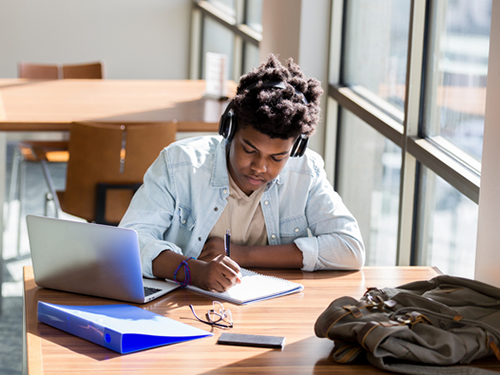 High school student studying in library, wearing headphones, working on laptop