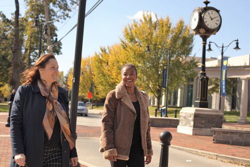 A female administrator walks on Widener's campus with a female student to her right. Widener's clock tower stands prominently in the background.