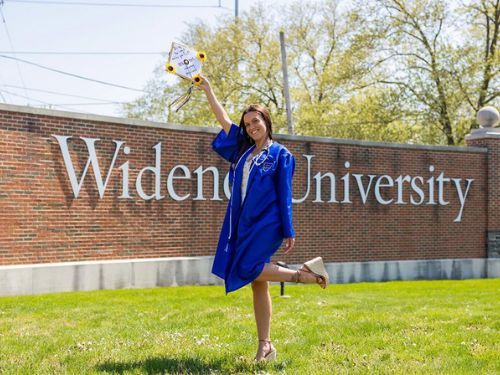 Female student celebrates in front of the Widener University sign with a cap, gown, and stethoscope around her neck.