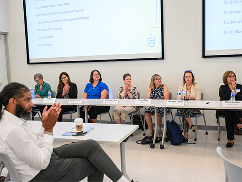 Seven professors sitting on a panel in front of a room, with an audience member clapping in the foreground