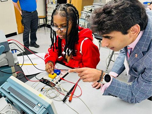 Professor Ali Hamza leans on a desk and points as a student works with a circuit