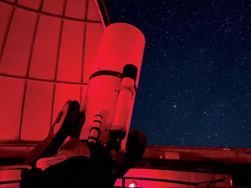 Telescope bathed in a red glow pointing out the open observatory toward a night sky full of stars