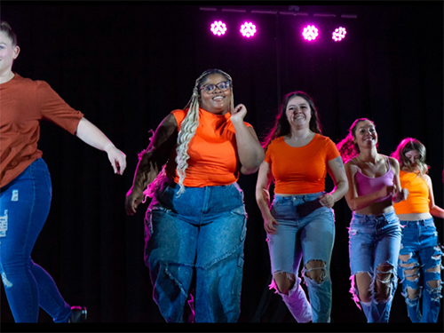 Ivana Brock, dancing on stage, among a group of other dances in matching jeans and orange shirts