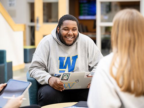 Justin Mack, wearing a Widener hoodie and holding a laptop, smiles as he talks to another student in the atrium of Quick Center