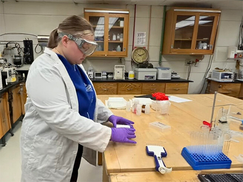 Engineering Student Bella Kuehn, wearing a white lab coat and protective goggles, works on material at a lab table