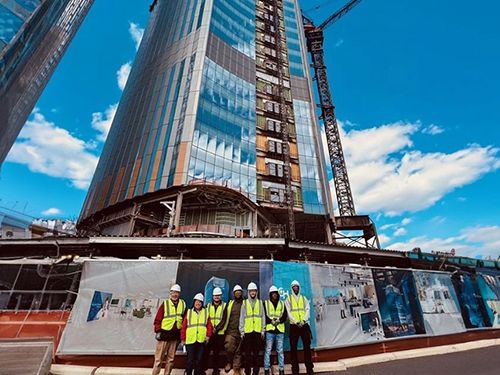 Seven students in hard hats and yellow construction vests stand in front of a building under construction