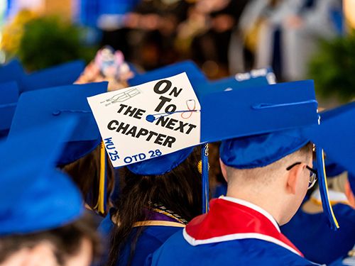 Back of graduates in blue caps and gowns with the cap in the center decorated in white with a picture of crutches and a stethoscope and the words “On to the next chapter. WU 25 OTD 28”