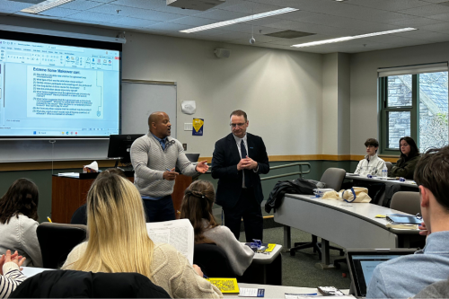 Deans Todd Clark and andré douglass pond cummings stand at the front of the room speaking to students. They have a presentation displayed on the screen behind them.