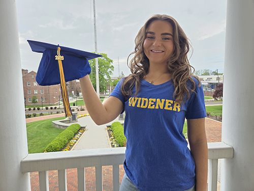 Amanda Rappa, wearing a blue Widener shirt and holding a blue grad cap, stands on the balcony of Old Main with campus behind her