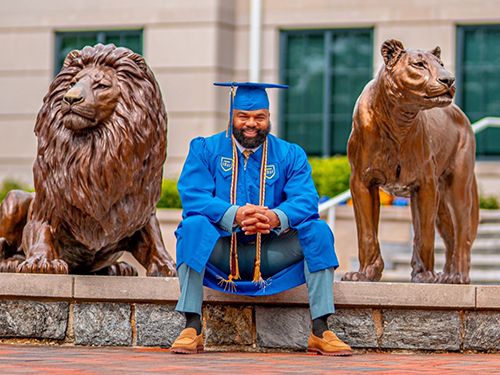 Delorean Andrews in blue cap and gown and cords sits between the Chester and Melrose lion statues