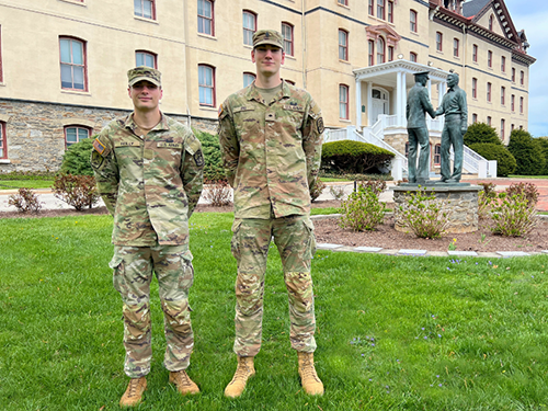R-O-T-C cadets Liam Reilly and Braydn Erb, wearing Army fatigues, stand on the grass in front of Old Main with a statue in the background