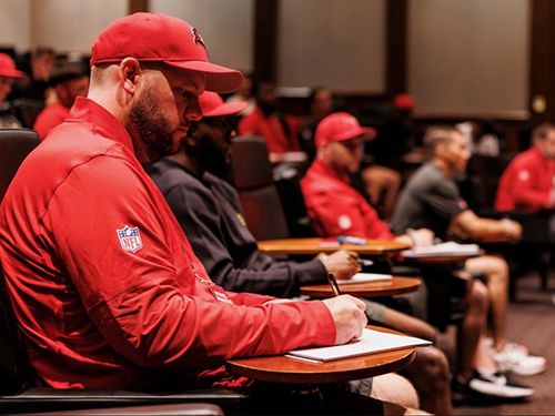 Mike Barainyak, wearing red NFL jacket and hat, sits at a desk taking notes