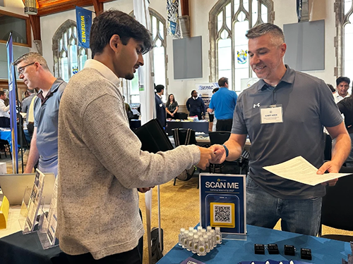 An employee at a career fair stands at a table and hands a student something while others mingle behind them
