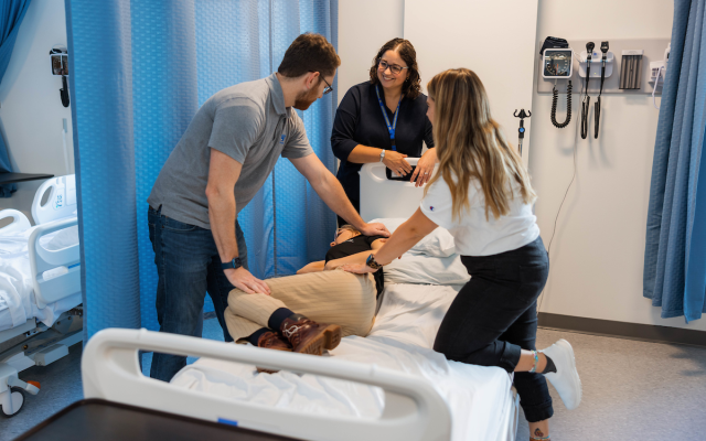 Two students helping to transfer a client out of a hospital bed, with a faculty member providing instruction