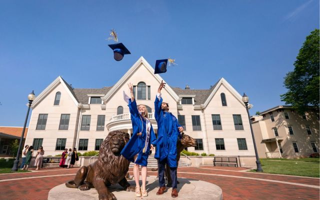 A female and male grad toss their caps in the air in front of the Pride statue.