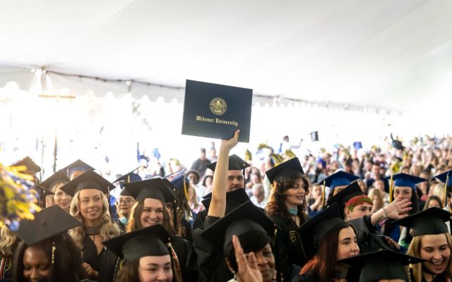 A graduate holds up a diploma holder that reads "Widener University" during a commencement ceremony under the tent.