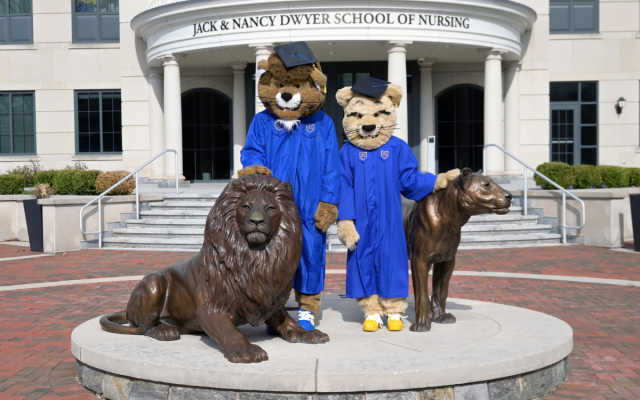 Widener mascots Chester and Melrose wear blue graduation caps and gowns and pose for a photo with the pride of lions statue