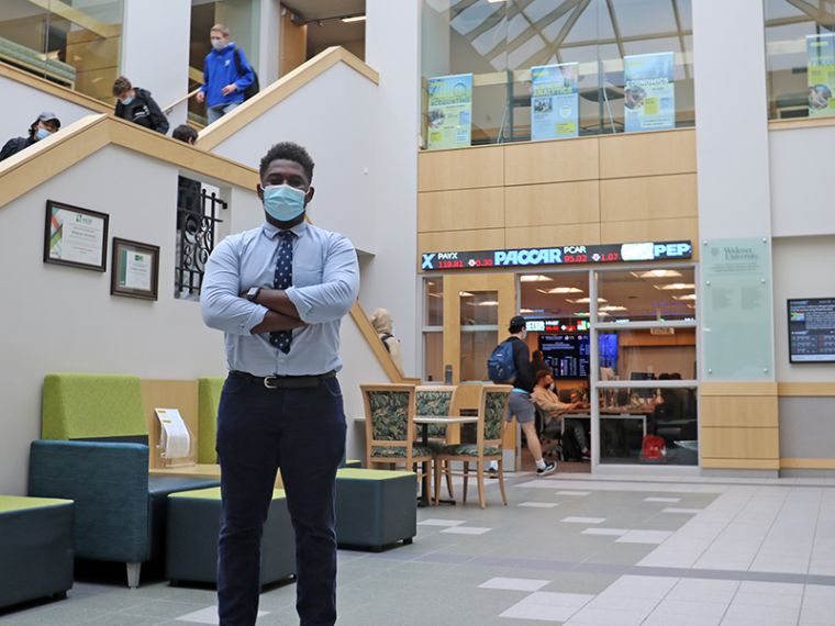 Brandon Matambo stands in the lobby of Quick Center; behind him is a stock ticker and students walking down steps