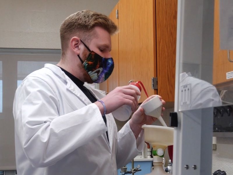 Aidan Looby, wearing a white lab coat and mask, works with equipment in a science lab