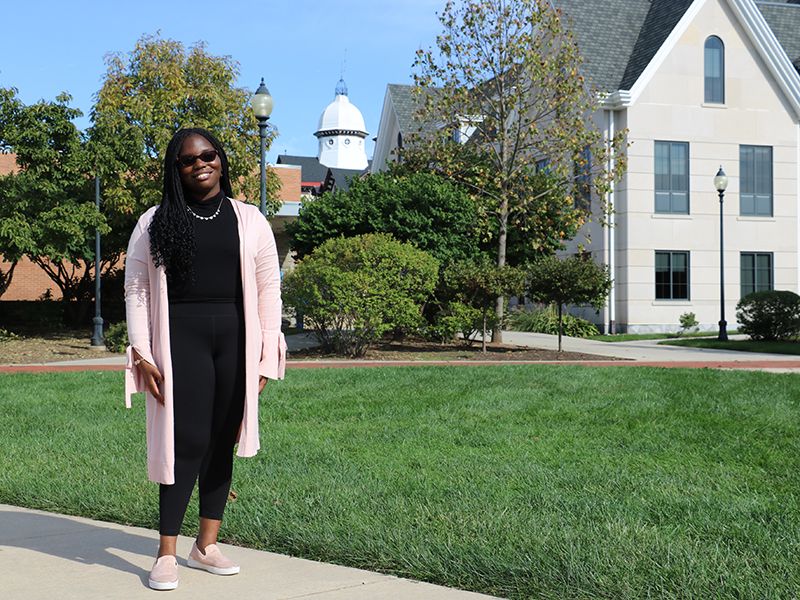 Kennedy Shaw stands on campus on a blue sky day, with Founders Hall behind her and the Old Main dome visible in the background